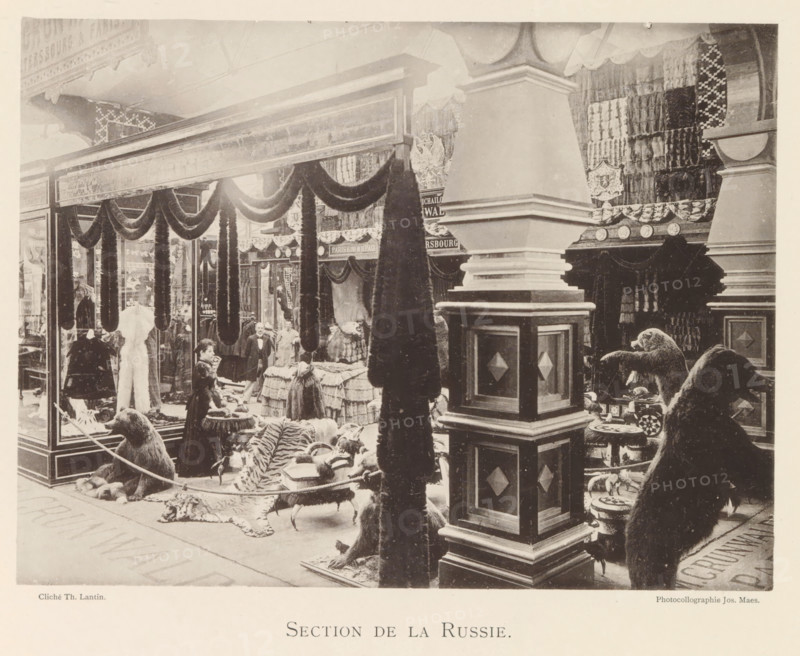 Section de la Russie, Exhibition of Russian fur products during the World Exhibition in Antwerp, page, photomechanical print, photographer: Th. Lantin, Antwerp, clichémaker: Joseph Maes, 1894, paper, collotype, photogravure, height 163 mm x width 219 mm, world fair, world exhibition, exhibition, show, furrier, A large indoor display area with tall decorative columns and draped fabric swags framing a storefront scene, shelves and racks filled with folded textiles and fur garments, several standing fur coats and hanging pelts, a woman near a counter arranging items, other figures standing behind tables covered in stacked goods, rugs and animal hides spread on the floor including a striped hide, mounted taxidermy bears positioned near stools and low tables, ornate carved wood and paneling on the pillars, patterned wall hangings and shields on the back walls, and a glass display case at the left showing garments inside.