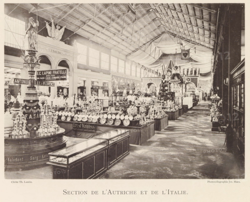 Exhibition of Austrian and Italian objects during the world exhibition in Antwerp, Section de l'Autriche et de l'Italie, page, photomechanical print, photographer: Th. Lantin, Antwerp, cliché maker: Joseph Maes, 1894, paper, collotype, light printing, height 160 mm x width 218 mm, world fair, world exhibition, exhibition, show, A wide interior view of a large exhibition hall with a high, trussed ceiling draped with fabric, rows of display tables filled with plates, ceramics, and decorative objects, and glass-topped cabinets in the foreground. The left side shows tall statues and an ornate columnar display topped with a figurine, with arched openings and large windows along the wall behind. The central area features densely arranged merchandise and a decorative pavilion structure with patterned panels and hanging banners, while the right side opens onto a long aisle with more displays and wall-mounted lights. The floor shows a wood texture and the overall scene is sepia-toned with stron...