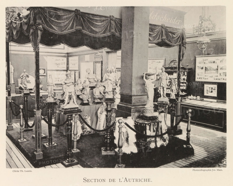 Section de l'Autriche, Exhibition of Austrian sculptures during the World Exhibition in Antwerp, page, photomechanical print, photographer: Th. Lantin, Antwerp, cliché maker: Joseph Maes, 1894, Th. Lantin, Joseph Maes, photographer, cliché maker, paper, collotype, photoprint, height 160 mm x width 220 mm, sculpture, world fair, world exhibition, exhibition, show, The image shows an indoor display area with a raised platform bordered by decorative posts and rope barriers, draped fabric canopies with tassels overhead, multiple carved statues and figurines arranged on pedestals and tables, a large central column, framed pictures on the walls, a chandelier hanging from the ceiling, and wooden floorboards visible around the exhibit.
