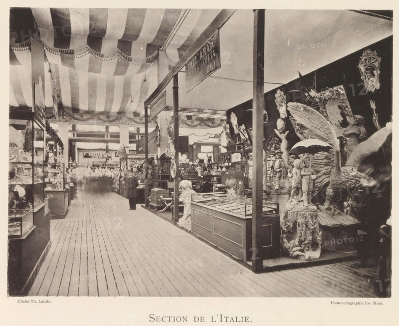 Section de l'Italie, Exhibition of Italian objects during the world exhibition in Antwerp, page, photomechanical print, photographer: Th. Lantin, Antwerp, cliché maker: Joseph Maes, 1894, Th. Lantin, Joseph Maes, photographer, cliché maker, paper, collotype, light printing, height 165 mm x width 220 mm, world fair, world exhibition, exhibition, show, A large indoor exhibition space with a wooden plank floor and a striped fabric ceiling draped across the top, supported by vertical posts that divide display areas; glass display cases and shelves line the left side and central aisles, while the right side features a dense arrangement of decorative sculptures and carved figures including a winged creature, a pair of child figures holding an umbrella, and various ornate frames and statues, all positioned on pedestals and platforms; a few blurred human figures are visible among the displays and a hanging rectangular sign is suspended from the ceiling structure.