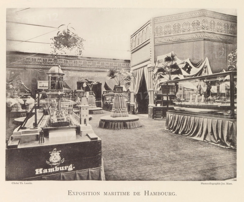 Exhibition of maritime objects from Hamburg during the World Exhibition in Antwerp, Exposition maritime de Hambourg, page, photomechanical print, photographer: Th. Lantin, Antwerp, clichémaker: Joseph Maes, 1894, paper, collotype, photogravure, height 163 mm x width 218 mm, ship-model, world fair, world exhibition, exhibition, show, A large indoor exhibition space with ornate patterned frieze along the upper walls and tall draped curtains dividing sections, containing several display cases and model maritime objects arranged on plinths and tables; a suspended hanging plant hangs near the ceiling and potted palm plants sit atop decorative round pedestals with skirted bases scattered through the room; a prominent model ship with rigging is positioned on a foreground table and other ship models are visible in glass cases and on stands; a corner structure with arched decorative panels rises at one side and fabric flags or cloths drape over part of the displays; wooden flooring covers the f...