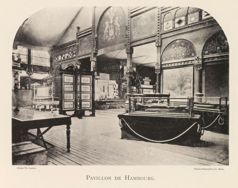 Interior of the pavilion from Hamburg during the World Exhibition in Antwerp, Pavillon de Hambourg, page, photomechanical print, photographer: Th. Lantin, Antwerp, clichémaker: Joseph Maes, 1894, paper, collotype, photogravure, height 160 mm x width 219 mm, interior, representation of a building, world fair, world exhibition, exhibition, show, A large interior space with a high curved ceiling and patterned decorative panels along the upper walls, featuring ornate arches and floral motifs. Wooden columns and carved supports rise from the floor to the decorated frieze, with a hanging fringed banner bearing a heraldic emblem. Display cases with glass tops sit on draped tables and pedestals, and a freestanding multi-paneled screen shows framed images. A wooden table with turned legs occupies the foreground, and ropes form a barrier around some exhibits. In the left background there is a raised platform with additional decorative furnishings and a tall window letting in light.