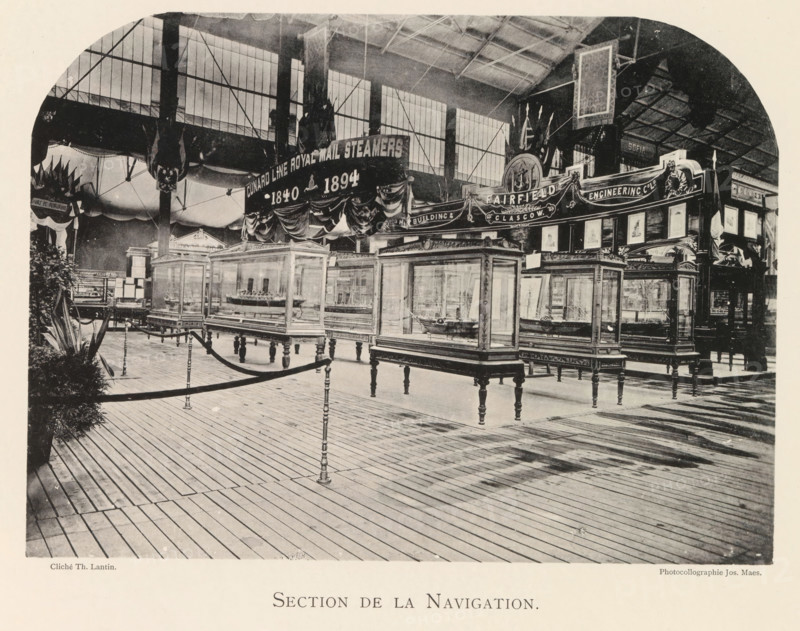 Exhibition of ship models during the World Exhibition in Antwerp, Section de la Navigation, page, photomechanical print, photographer: Th. Lantin, Antwerp, clichémaker: Joseph Maes, 1894, paper, collotype, photoprint, height 160 mm x width 218 mm, ship-model, world fair, world exhibition, exhibition, show, An indoor exhibition space with a high roof and large windows shows rows of glass display cases on ornate wooden legs containing model ships, a wooden plank floor with rope barriers supported by decorative posts, draped banners and decorative panels above the displays, tall vertical supports and hanging decorative elements, framed pictures and ornamental signage along the back, and potted plants visible at the left edge.