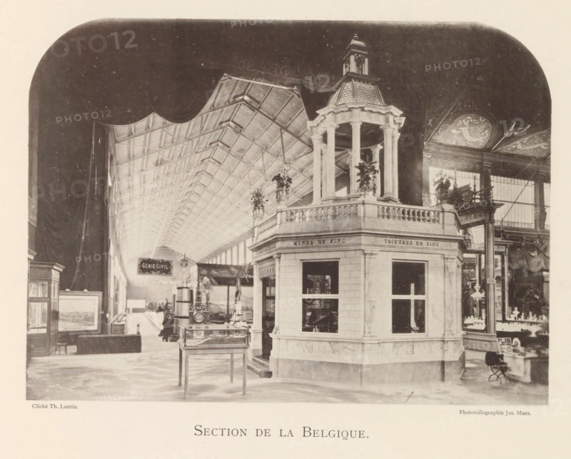 Exhibition of Belgian objects during the World Exhibition in Antwerp, Section de Belgique, page, photomechanical print, photographer: Th. Lantin, Antwerp, clichémaker: Joseph Maes, 1894, paper, collotype, photoprint, height 160 mm x width 220 mm, world fair, world exhibition, exhibition, show, A large interior space with a high vaulted ceiling composed of repeating trusses extends into the distance, framed by a rounded dark border at the top edges. In the foreground a multi-sided pavilion with balustraded upper railings and tall columns supporting a small domed cupola stands prominently, with hanging plants placed near its corners. Glass display cases and framed artworks are arranged along the floor area, and a few freestanding decorative streetlamp-like structures rise near the pavilion. Ornate painted panels with floral and bird motifs decorate a wall to the right, and tall windows admit light behind the decorative elements. Various small objects and fixtures are visible on tables an...