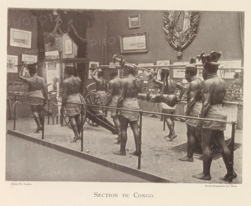 Sculptures in the Congolese part of the world exhibition in Antwerp, Section du Congo, page, photomechanical print, photographer: Th. Lantin, Antwerp, cliché maker: Joseph Maes, 1894, paper, collotype, photoprint, height 160 mm x width 219 mm, sculpture, world fair, world exhibition, exhibition, show, A sepia-toned scene showing a museum-like display of several bare-chested men wearing wrapped cloths around their waists and standing barefoot on a sandy floor, carrying a long pole with a mounted cylindrical object and wagon wheel parts, with framed paintings and decorative shields hung on the wall behind them, a roped barrier in front of the display, and a few additional artifacts and small objects arranged on tables along the back wall.