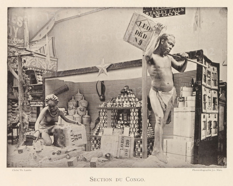 Sculptures in the Congolese part of the world exhibition in Antwerp, Section du Congo, page, photomechanical print, photographer: Th. Lantin, Antwerp, cliché maker: Joseph Maes, 1894, paper, collotype, photoprint, height 160 mm x width 218 mm, sculpture, world fair, world exhibition, exhibition, show, A sepia-toned photograph showing two partially clothed individuals posed among stacked boxes, jars, woven baskets, and various packaged goods on a display area framed by decorative panels and a star ornament; a seated figure wearing a beaded necklace and a cloth around the waist is reaching toward containers on the ground while a standing figure balanced on one leg holds a wooden pole across the shoulders and a crate over one shoulder; behind them are shelves filled with stacked jars and cylindrical containers, bundled brooms or reeds on the floor, and a tall display cabinet with panels showing relief images.