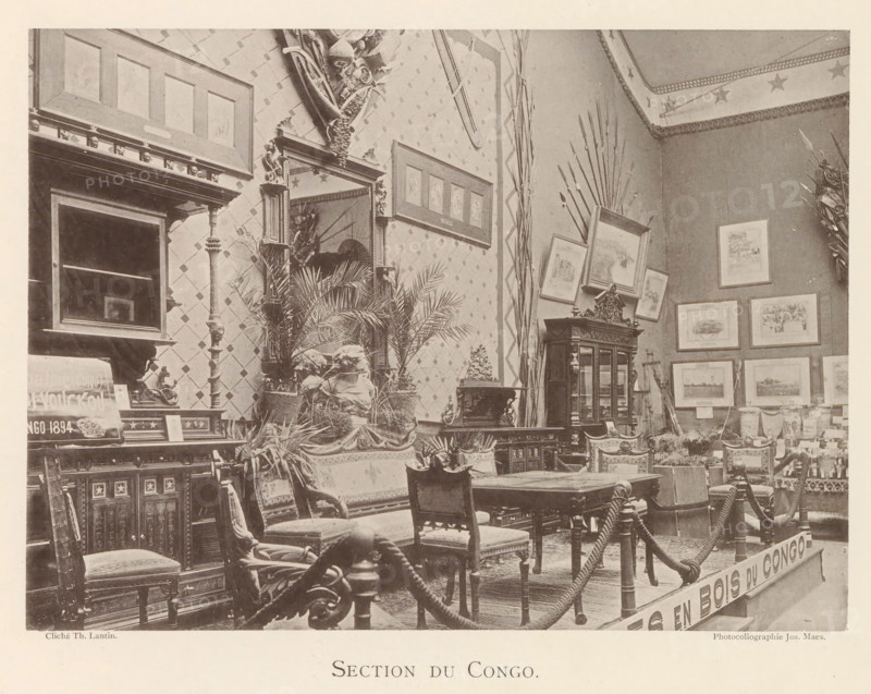 Exhibition of wooden furniture from Congo at the world exhibition in Antwerp, Section du Congo, page, photomechanical print, photographer: Th. Lantin, Antwerp, cliché maker: Joseph Maes, 1894, paper, collotype, light printing, height 160 mm x width 219 mm, world fair, world exhibition, exhibition, show, seating furniture, A room filled with ornate wooden furniture including carved chairs, a table, cabinets, and a large mirrored mantle, surrounded by framed pictures on patterned walls and decorative spears leaning against the corner, with potted plants and sculpted busts arranged on and around the furniture, a rope barrier suspended between posts in the foreground, and a ceiling border decorated with star motifs.