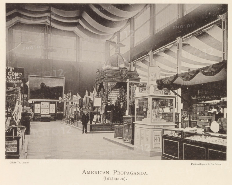 Interior of the American pavilion at the World's Fair in Antwerp, American propaganda (interior), page, photomechanical print, photographer: Th. Lantin, Antwerp, printer (person): Joseph Maes, 1894, Th. Lantin, Joseph Maes, photographer, printer (person), paper, collotype, photogravure, height 153 mm x width 219 mm, interior, representation of a building, world fair, world exhibition, exhibition, show, A large indoor exhibition hall with a high ceiling draped in wide scalloped fabric panels and tall windows along the upper wall, a central ornate display topped with a domed structure and an eagle figure, several men in period clothing standing on and around the raised platform in front of the display, a uniformed man walking on the main wooden floor, various booths and counters filled with merchandise and display cases to the right and left, decorative flags and framed artwork against the far wall, hanging awnings and fabric swags above some booths, and wooden flooring stretching throug...