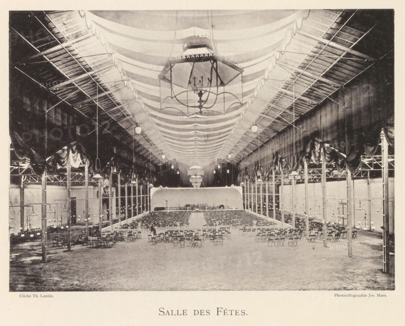 Interior of a banquet hall in Antwerp, Salle des fêtes, page, photomechanical print, photographer: Th. Lantin, Antwerp, printer (person): Joseph Maes, 1894, printer (person), paper, collotype, photoprint, height 160 mm x width 219 mm, public festivities, interior, representation of a building, A large indoor hall with a high vaulted ceiling draped with fabric swags and a central oversized chandelier suspended above rows of folding chairs arranged facing a raised platform at the far end; tall slender columns line both sides supporting decorative drapery and overhead trusses, with hanging globe lights spaced along the ceiling and smaller chandeliers mounted on poles near the aisles, while the floor appears bare and expansive in the foreground.