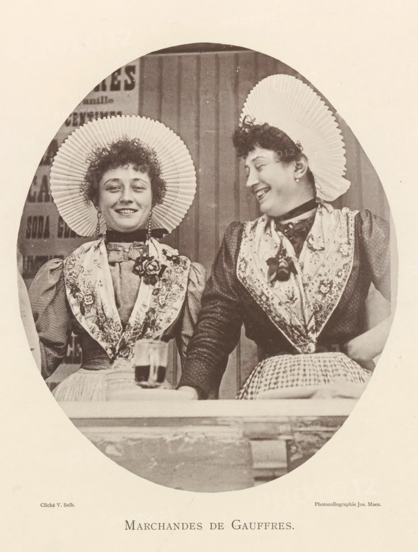 Portrait of two waffle sellers in Antwerp, Marchandes de gauffres, page, photomechanical print, photographer: Victor Selb, Antwerp, printer (person): Joseph Maes, c. 1890 - in or before 1894, printer (person), paper, collotype, photoprint, height 203 mm x width 172 mm, merchant, salesman - BB - women engaged in trade and commerce, waffles, Two women sit side by side inside an oval frame, both wearing pleated round hats and patterned shawls over high-collared dresses; the woman on the left faces forward with a slight smile while the woman on the right turns toward her with a broader smile, and both have short curly hair and earrings. A table edge is visible in front of them with a glass placed on it, and the background shows vertical wooden panels. The image is sepia-toned.