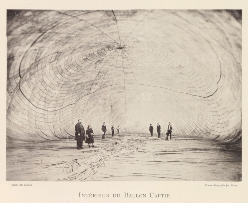 Intérieur du Ballon Captif, People in a tethered balloon during the world exhibition in Antwerp, page, photomechanical print, photographer: Th. Lantin, Antwerp, cliché maker: Joseph Maes, 1894, paper, collotype, photoprint, height 159 mm x width 217 mm, balloon (aviation), world fair, world exhibition, exhibition, show, A large tunnel-like interior with a curved, ribbed surface stretching overhead and inward toward a distant center, the walls marked by concentric arc patterns and subtle shading variations; a textured floor with folds and creases runs toward the center of the space; several people stand along the floor at varying distances from the foreground, wearing long coats and period clothing and facing toward the camera, some holding objects or with hands at their sides, their features and postures visible against the pale interior backdrop.