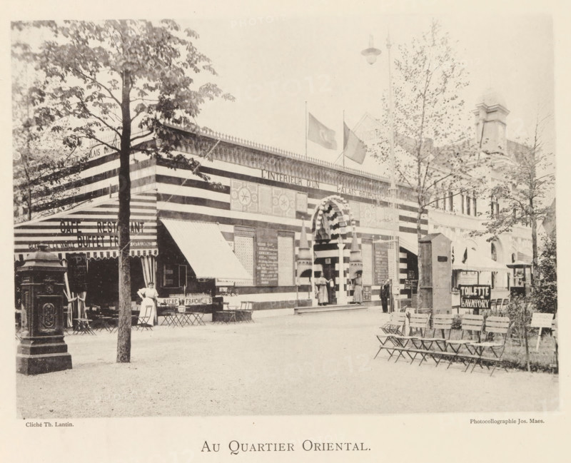 At the Oriental Quarter, Exterior of the Oriental section of the World Exhibition in Antwerp, page, photomechanical print, photographer: Th. Lantin, Antwerp, printer (person): Joseph Maes, 1894, printer (person), paper, collotype, photogravure, height 160 mm x width 219 mm, façade (of house or building), world fair, world exhibition, A wide exterior scene shows a striped building facade with an ornate arched entrance flanked by columns and decorative panels, flags flying above the roofline, and patterned awnings extending from the front; several people stand near the entrance and a woman in a dress stands by a cluster of small tables and chairs on the left; trees with leaves frame parts of the scene and a row of folding chairs lines the right foreground; a freestanding ornamental structure stands near the left edge and a tall lamp hangs above the street.