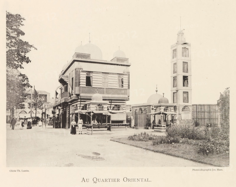 Exterior of the Oriental section of the World Exhibition in Antwerp, Au quartier oriental, page, photomechanical print, photographer: Th. Lantin, Antwerp, printer (person): Joseph Maes, 1894, printer (person), paper, collotype, photogravure, height 155 mm x width 220 mm, façade (of house or building), world fair, world exhibition, A street scene showing a large multi-story building with striped exterior walls, decorative latticework, and domed roof elements, adjacent to a tall square tower with open lattice panels and a narrow spire; market stalls with awnings and counters line the front of the building where a few people stand and a person sits behind a counter; to the left, more pedestrians walk along the tree-lined street and smaller buildings are visible in the background; to the right, a fenced area with small domed structures, decorative entrances, and garden beds occupies the foreground.
