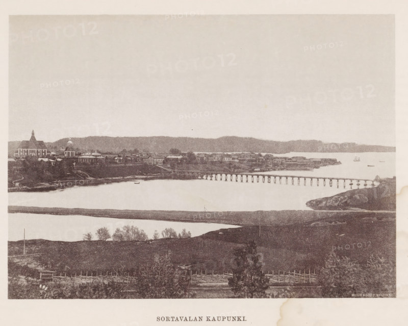Sortavalan Kaupunki, View of Sortavala, page, photomechanical print, photographer: Impi Backman, Sortavala, cliché maker: anonymous, printer (person): Weilin & Göös, c. 1888 - c. 1893, printer (person), paper, autotypie, height 163 mm x width 224 mm, Print two pages for page 1., city-view in general, 'veduta', landscapes with waters, waterscapes, seascapes (in the temperate zone), bridge, A wide sepia-toned panorama shows a shoreline town across a calm body of water with a long wooden pier stretching from the right toward the center; buildings with varied rooflines and a few domed and spired structures sit along the distant shore backed by a low ridge of land, and a small sailboat is visible on the water; in the foreground there are grassy and rocky fields with scattered trees and simple wooden fences, and a strip of land or sandbar runs parallel to the nearer shore.
