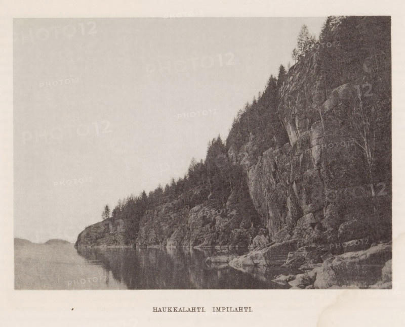 Rocks by a water, Haukkalahti. impilahti, page, photomechanical print, photographer: Impi Backman, cliché maker: anonymous, printer (person): Weilin & Göös, c. 1888 - c. 1893, printer (person), paper, autotypie, height 153 mm x width 212 mm, trees, landscapes with waters, waterscapes, seascapes (in the temperate zone), rocks, Karelia, A monochrome photograph showing a rocky shoreline with steep cliffs rising from calm water, trees growing along the cliff tops and scattered on rocky ledges, reflections of the rocks and trees visible in the smooth surface of the water, and distant low hills fading into the background under a pale sky.