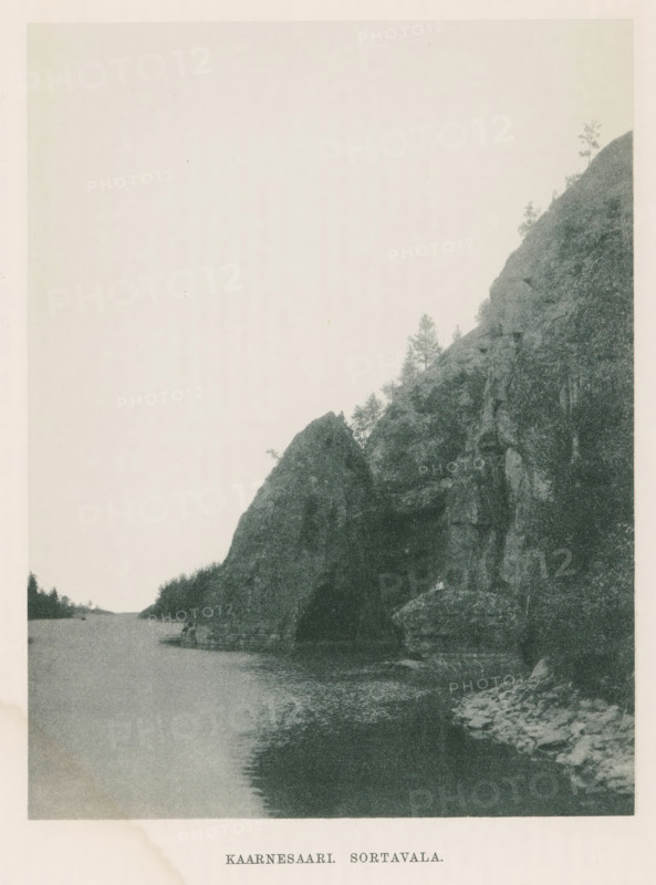 Kaarnesaari. Sortavala, Rocks on a water, page, photomechanical print, photographer: Impi Backman, cliché maker: anonymous, printer (person): Weilin & Göös, c. 1888 - c. 1893, printer (person), paper, autotypie, height 224 mm x width 163 mm, Print three pages after page 24., trees, landscapes with waters, waterscapes, seascapes (in the temperate zone), rocks, Karelia, A vertical photograph showing a calm body of water along a rocky shoreline with steep cliffs rising on the right side; the cliffs have rough textured rock faces and scattered small trees and shrubs growing on ledges and near the top; a large dark rock formation juts into the water creating a shadowed area along the shore; the water surface is smooth with faint ripples reflecting the dark tones of the rocks; a narrow strip of distant shoreline is visible on the left with low vegetation.