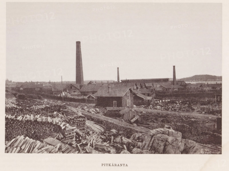 Chimneys near buildings and stacks of wood, Pitkäranta, page, photomechanical print, photographer: Impi Backman, cliché maker: anonymous, printer (person): Weilin & Göös, c. 1888 - c. 1893, printer (person), paper, autotypie, height 157 mm x width 226 mm, civic architecture, edifices, dwellings, chimney, industries, plant material, A sepia-toned landscape showing a shoreline area with rocky foreground and scattered piles of cut logs and planks, a cluster of simple wooden houses and outbuildings with pitched roofs set behind the rocky area, a larger industrial complex behind the houses with long low buildings and several tall narrow smokestacks rising into the pale sky, distant low hills and water visible along the horizon, and an expansive empty sky occupying the upper portion of the image.
