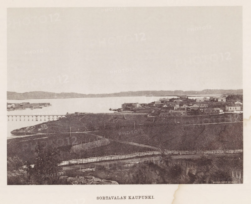 View of Sortavala, Sortavalan Kaupunki, page, photomechanical print, photographer: Impi Backman, Sortavala, cliché maker: anonymous, printer (person): Weilin & Göös, c. 1888 - c. 1893, printer (person), paper, autotypie, height 157 mm x width 210 mm, city-view in general, 'veduta', landscapes with waters, waterscapes, seascapes (in the temperate zone), bridge, A wide sepia-toned shoreline view with a calm body of water stretching across the middle, a distant low ridge on the far side, and a small cluster of buildings and houses along the near shore including piers and docks; in the foreground there are fenced plots and narrow paths running across a grassy, uneven landscape with rocks and scattered vegetation, and a faint sky occupying the upper portion of the image.
