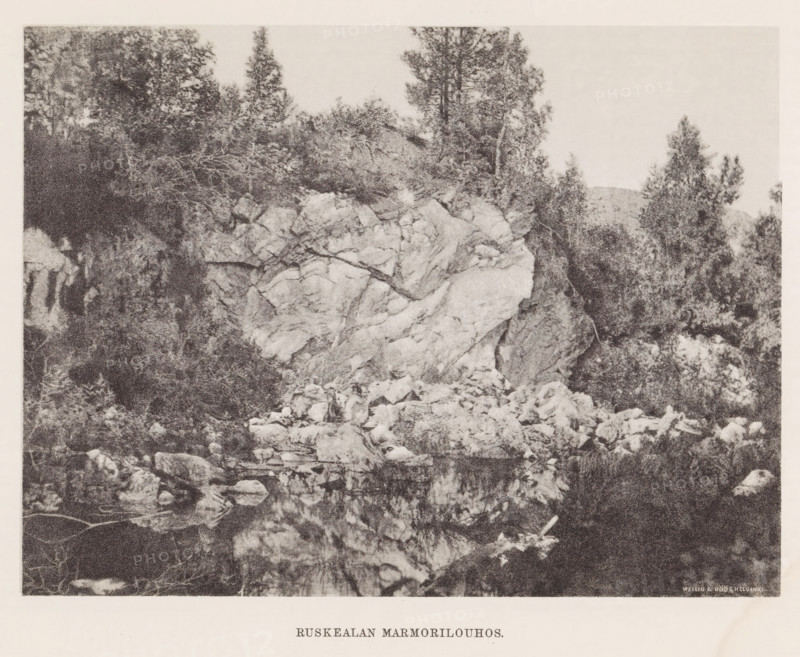 View of rocks by a body of water, Hämekoski. Ruskeala, page, photomechanical print, photographer: Impi Backman, cliché maker: anonymous, printer (person): Weilin & Göös, c. 1888 - c. 1893, printer (person), paper, autotypie, height 168 mm x width 225 mm, landscapes with waters, waterscapes, seascapes (in the temperate zone), rocks, Karelia, A grainy black-and-white photograph showing a rocky cliff face with large jagged boulders at its base and a reflective body of water in front, dense clusters of trees and shrubs growing atop and around the rock formation, a variety of tree shapes including tall slender trunks and bushier foliage, scattered smaller rocks and debris along the shoreline, and a clear sky above.