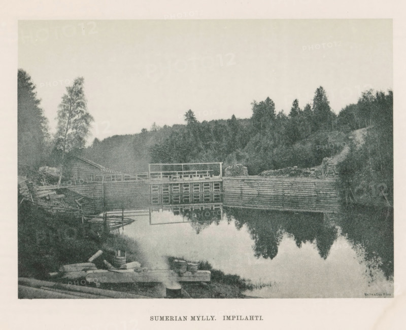 Sumerian Mylly. Impilahti, View of waterworks in a river, page, photomechanical print, photographer: Impi Backman, cliché maker: anonymous, printer (person): Weilin & Göös, c. 1888 - c. 1893, printer (person), paper, autotypie, height 161 mm x width 216 mm, trees, landscapes with waters, waterscapes, seascapes (in the temperate zone), Karelia, A sepia-toned riverside scene showing a wooden dam structure across calm water with its reflection visible, a dock and small boats moored near the bank, stacked logs and barrels on the shore, a simple wooden building and fences near the water, dense trees and foliage rising behind the structures, and a pale sky above.