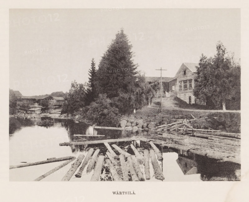 View of tree trunks in the water and houses around the water, Wärtsilä, page, photomechanical print, photographer: Impi Backman, cliché maker: anonymous, printer (person): Weilin & Göös, c. 1888 - c. 1893, printer (person), paper, autotypie, height 166 mm x width 222 mm, landscapes with waters, waterscapes, seascapes (in the temperate zone), civic architecture, edifices, dwellings, Karelia, A riverside scene showing a calm body of water with floating logs and a simple wooden raft in the foreground, a pile of timber and scattered branches along the shore, a rocky bank leading up to a dirt path, tall evergreen trees and other dense foliage rising on the riverbank, a wooden building with a pitched roof partially visible behind the trees, a simple wooden cross mounted near the path, and a treeline and additional low buildings visible across the water.