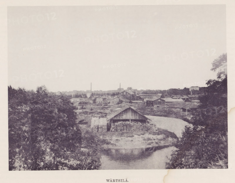 Wood storage facility in a landscape, Wärtsilä, page, photomechanical print, photographer: Impi Backman, cliché maker: anonymous, printer (person): Weilin & Göös, c. 1888 - c. 1893, printer (person), paper, autotypie, height 164 mm x width 217 mm, landscapes with waters, waterscapes, seascapes (in the temperate zone), civic architecture, edifices, dwellings, Pohjois-Karjala, Värtsilä, Vyartsilya, A grainy monochrome scene showing a riverside with several wooden buildings and sheds along the bank, a larger barn-like structure near the center with stacks of firewood visible under its overhang, a narrow waterway curving past the foreground reflecting surrounding shapes, dense trees and foliage framing both sides of the image, a low distant skyline of more industrial and residential buildings and two tall slender chimneys rising above the roofs, and a wide pale sky occupying the upper portion of the picture.