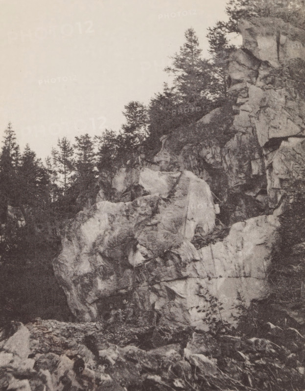 Ruskaelan Marmorilouhos, Rocks in Ruskeala, page, photomechanical print, photographer: Impi Backman, cliché maker: anonymous, printer (person): Weilin & Göös, c. 1888 - c. 1893, printer (person), paper, autotypie, height 215 mm x width 167 mm, rocks, Sortavala, A tall rocky cliff face occupies the right side of the image, showing layered rough stone with visible cracks and ledges, while trees and dense foliage grow along the top edge and in the background on the left; the foreground contains scattered rocks and boulders at the cliff base, and the sky above is clear and light in tone.
