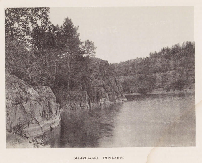 View of a river, Majatsalmi Impilahti, page, photomechanical print, photographer: Impi Backman, cliché maker: anonymous, printer (person): Weilin & Göös, c. 1888 - c. 1893, printer (person), paper, autotypie, height 152 mm x width 206 mm, trees, river, rocks, Karelia, A wide monochrome scene showing a rocky shoreline with steep cliffs and rough textured rock faces rising from calm water, trees and dense forest vegetation covering the cliff tops and extending into the background, a smooth expanse of water reflecting subtle ripples along the shore, and a pale sky above with no visible clouds.