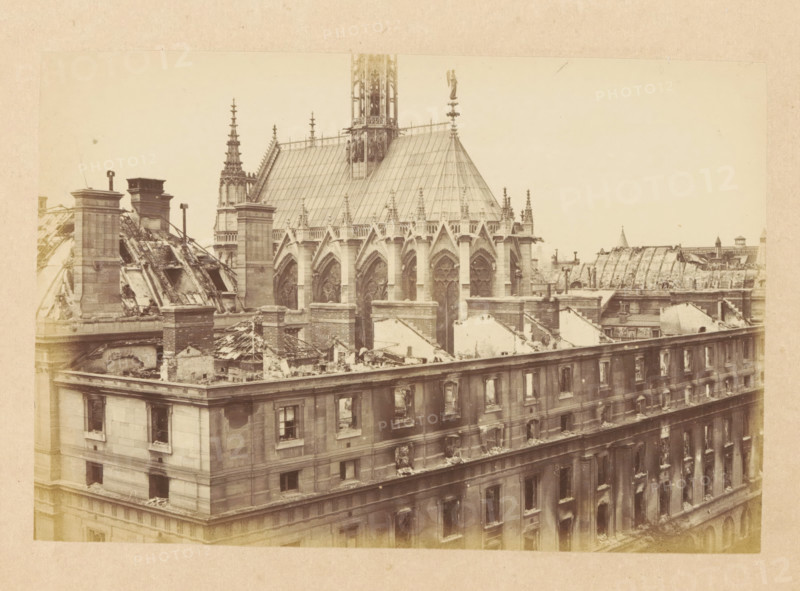 View of a destroyed Palace of Justice in Paris, Ensemble du Palais de Justice, photograph, page, 1871, photographer, paper, albumen print, albumendruk, height 101 mm x width 151 mm, ruin of a dwelling, house, castle, palace, Paris, Palais de Justice, A sepia-toned photograph shows the upper facade and roofline of a large stone building with Gothic architectural elements, including pointed arches and ornate tracery on tall windows beneath a steeply pitched roof with decorative pinnacles and a slender spire. The roof and parts of the upper walls appear damaged, with exposed beams, rubble, and partial collapses revealing interior spaces and broken masonry. Chimneys and rooftop structures rise above the ruined sections, and the row of rectangular windows on the building's lower stories shows soot-darkened frames and broken panes. In the background, additional rooftops and architectural details are visible against a pale sky.