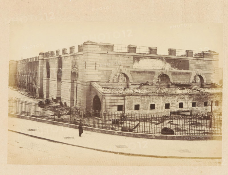 Grenier d'Abondance - vue extérieure, View of a destroyed building, photograph, page, 1871, photographer, paper, albumen print, albumendruk, height 101 mm x width 151 mm, ruin of a dwelling, house, castle, palace, Paris, A sepia-toned photograph of a large stone building with a long facade featuring arched openings and rectangular blocks of masonry, a lower section with small square openings and a roof edge showing debris, upper walls with arched window openings and some metal grilles, and a parapet with short square pillars and railings; in front of the building a fenced yard contains scattered rubble and large cylindrical objects, the fence running along a paved street where a solitary figure wearing a long coat and hat stands near the curb.