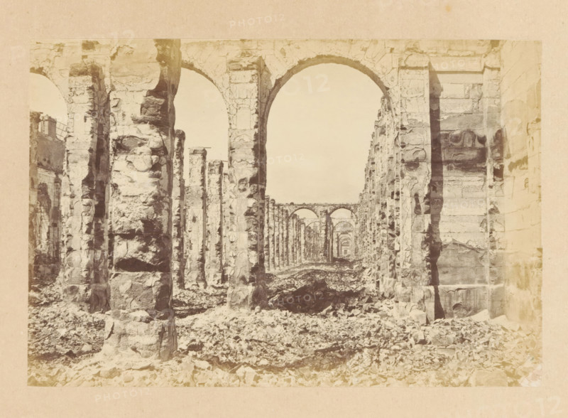 Grenier d'Abondance - vue intérieure, View of the interior of a destroyed building, photograph, page, 1871, photographer, paper, albumen print, albumendruk, height 101 mm x width 151 mm, ruin of a dwelling, house, castle, palace, Paris, A sepia-toned photograph of a long colonnade of stone arches and pillars seen from ground level, with rough, weathered stone surfaces and fractured edges, rubble scattered across the floor, light filtering through the open archways, multiple repeating arches receding into the distance, large vertical stone supports on either side, and a narrow view down the central aisle framed by the tall curved arch in the foreground.