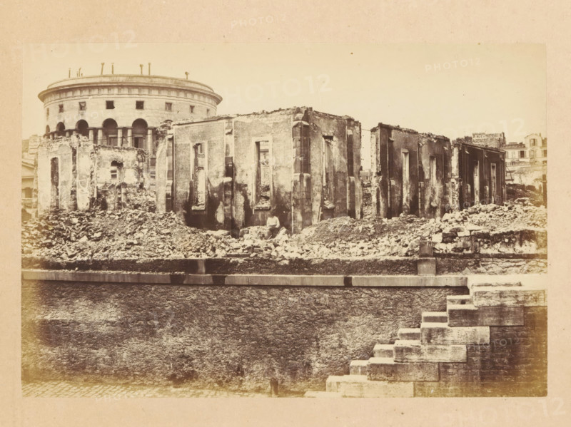 Docks de la Vilette, View of a destroyed building, photograph, page, 1871, photographer, paper, albumen print, albumendruk, height 101 mm x width 151 mm, ruin of a dwelling, house, castle, palace, A sepia photograph showing a row of damaged stone buildings with large rectangular window openings and rough, crumbling facades, piles of rubble and debris scattered along their bases, and a rounded multi-story structure with arched openings and a colonnade visible behind them, a low stone wall running across the foreground, and a stepped stone stairway descending at the right edge of the image.