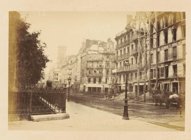View of destroyed buildings on Rue St Martin, Rue St Martin, photograph, page, Paris, 1871, photographer, paper, albumen print, albumendruk, height 101 mm x width 151 mm, street, theater (building), A sepia-toned street scene showing a row of multi-story buildings with visible damage and scaffolding against a pale sky, ornate wrought-iron balconies and shuttered windows on the facades, piles of debris and wooden supports at the bases of some buildings, a tall ornate street lamp in the foreground and another further down the street, a decorative iron fence and leafy trees along the left side, a domed kiosk set back near the buildings, and a horse-drawn cart partially visible at the lower right.