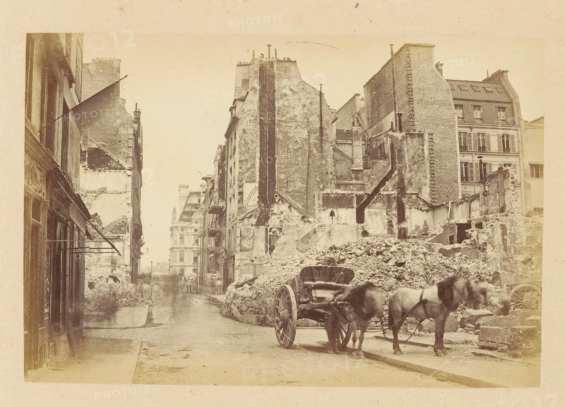Rue du Bac & Rue de Lille, View of a horse cart in front of destroyed buildings the Rue du Bac and the Rue de Lille, photograph, page, 1871, photographer, paper, albumen print, albumendruk, height 101 mm x width 151 mm, street, two-wheeled, animal-drawn vehicle, theater (building), Paris, A narrow urban street flanked by tall, weathered buildings with exposed brick and plaster, many showing damaged or partially demolished facades, rubble piled along the right side of the street, a wooden cart with large spoked wheels hitched to two horses standing near the rubble, a few indistinct figures further down the street, awnings and shopfronts visible on the left, remnants of interior walls and staircases visible in the ruined structures, and a distant multi-story building seen at the end of the street.