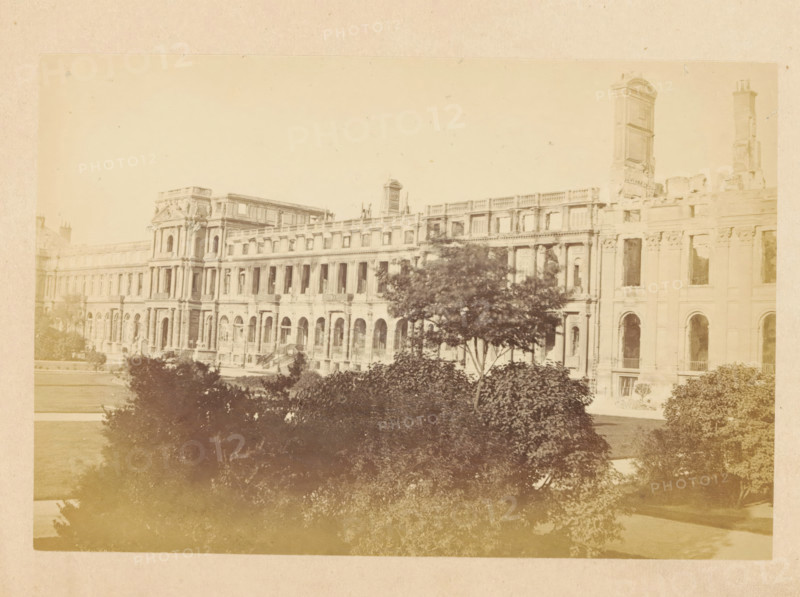 View of the Tuileries Palace in Paris, Palais des Tuileries - vue extérieure, photograph, page, Tuileries Palace, 1871, photographer, Paris, paper, albumen print, height 101 mm x width 149 mm, front facade, palace, A wide sepia-toned photograph of a large classical building with rows of tall arched windows and ornate columns, showing a long facade with multiple stories and decorative stonework, a partially ruined tower rising above the right side, and trees and shrubbery in the foreground partially obscuring the lower part of the building.
