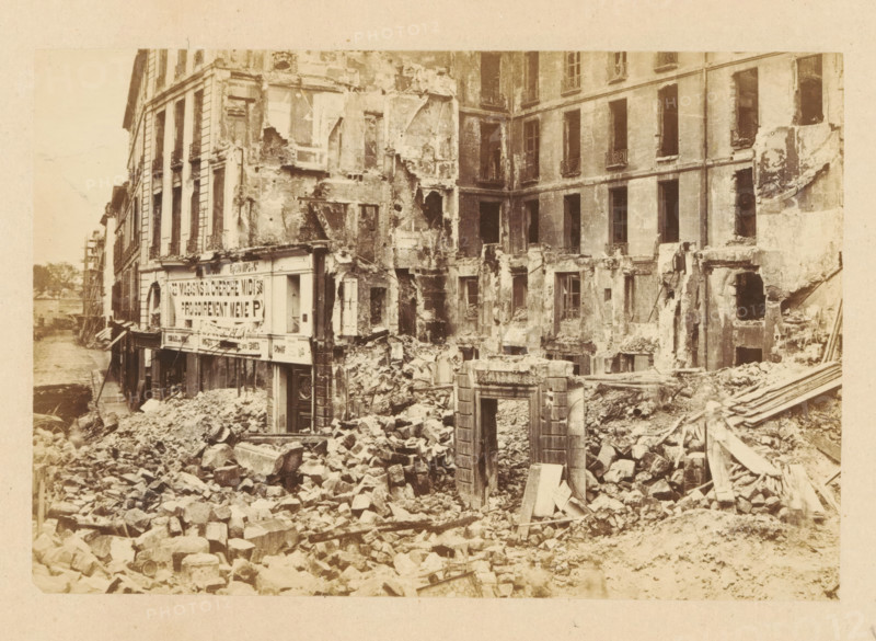 Carrefour de la Croix Rouge, View of a destroyed building at Carrefour de la Croix Rouge, photograph, page, 1871, photographer, paper, albumen print, albumendruk, height 101 mm x width 151 mm, street, theater (building), Paris, A sepia-toned scene of a heavily damaged multi-story building with large sections of facade missing and exposed interior walls, empty window openings, and crumbling plaster. The ground in front of the building is covered in a chaotic pile of rubble, broken stones, and timber debris. A partially intact doorway frame stands amid the debris, and fragments of architectural details lie scattered. Adjacent buildings show worn exteriors and shuttered or open windows, while a narrow street or waterway runs alongside the row of structures. The overall appearance is one of widespread structural collapse and scattered building materials.