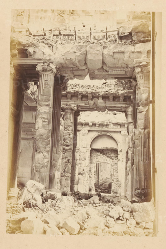 View of the ruined interior of the Tuileries Palace in Paris, Palais des Tuileries - vue interiore, photograph, page, Tuileries Palace, 1871, photographer, Paris, paper, albumen print, albumendruk, height 150 mm x width 101 mm, ruin of a dwelling, house, castle, palace, A sepia-toned view of a ruined stone interior showing tall fluted columns with ornate capitals, large blocks and broken masonry piled on the floor, a partially collapsed stone lintel overhead with visible carved cornices, fragments of decorative molding along the upper wall, a recessed archway in the background leading to another damaged opening, scattered rubble filling the foreground, and worn, cracked stone surfaces throughout.