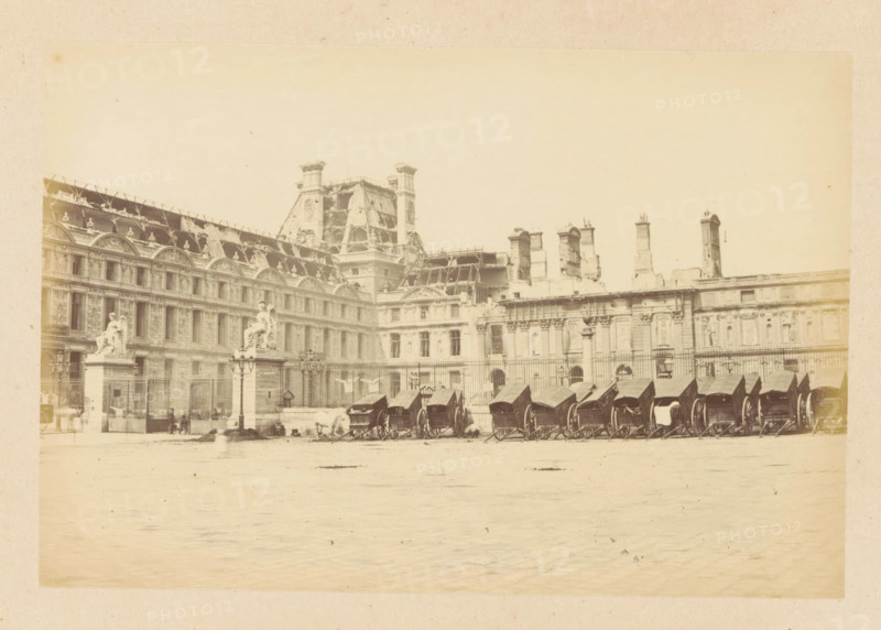 Palais des Tuileries - vue extérieurre, View of the destroyed Tuileries Palace in Paris, photograph, page, Tuileries Palace, 1871, photographer, Paris, paper, albumen print, height 101 mm x width 150 mm, ruin of a dwelling, house, castle, palace, A wide pale photograph of an ornate stone building with rows of arched windows and decorative roofline elements, including sculpted figures and ornamental pediments, a central section with a steeply pitched roof and scaffolding or exposed framework, and several tall chimneys and vertical architectural features along the roof. In front of the building a fenced courtyard contains a series of covered horse-drawn carriages lined up side by side, lamp posts, and two large sculpted statues mounted on tall pedestals near the entrance. The foreground shows a broad open paved area with faint shadows and some indistinct figures and blurs suggesting movement.