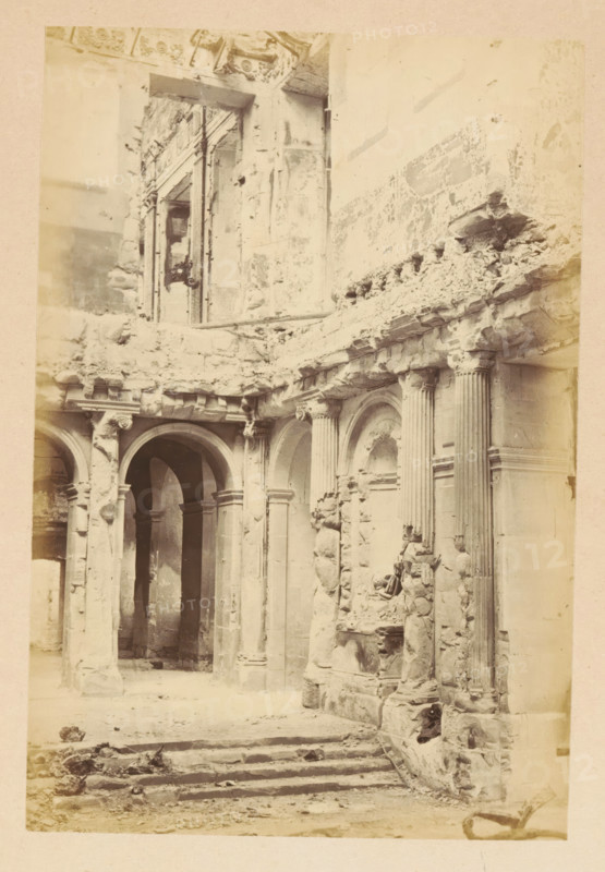 View of the ruined interior of the Tuileries Palace in Paris, Palais des Tuileries - vue intérieure, photograph, page, Tuileries Palace, 1871, photographer, Paris, paper, albumen print, albumendruk, height 150 mm x width 101 mm, ruin of a dwelling, house, castle, palace, A sepia-toned photograph of a ruined stone interior showing arched doorways and fluted columns with damaged capitals and bases, crumbling masonry and exposed rubble along the upper ledges, carved niches and ornamental stonework partly broken away, a set of worn stone steps leading up into the space, and fragments of stone and debris scattered across the floor.