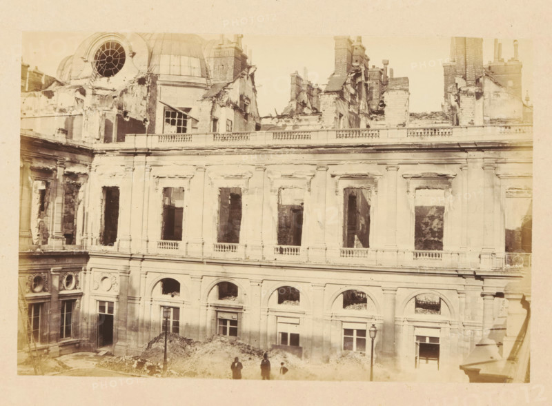 Palais Royal, View of the ruined Palais Royal, photograph, page, Palais-Royal, 1871, photographer, Paris, paper, albumen print, albumendruk, height 101 mm x width 151 mm, ruin of a dwelling, house, castle, palace, A sepia-toned photograph of a large multi-story stone building with classical architectural details showing extensive damage and missing windowpanes, with an upper section where rooflines and chimneys are jagged and partially collapsed, a circular window opening near a dome, decorative columns and balustrades along the facade, several arched openings on the lower level and tall rectangular window openings above, rubble piled against the base of the building, a single street lamp in front of the facade, and four small human figures standing near the rubble.