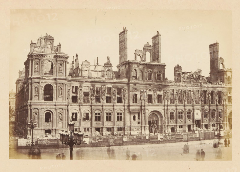 City Hall, View of the ruined Paris City Hall, photograph, page, 1871, photographer, Paris, paper, albumen print, height 101 mm x width 151 mm, ruin of a dwelling, house, castle, palace, A large stone building facade with ornate architectural details showing extensive damage and missing sections, multiple tall rectangular towers and partial walls rising above the roofline, rows of arched windows and niches with sculpted figures, broken openings and rubble along the base, scaffolding or temporary supports propped at a main arched entrance, a low fence or hoarding running along the front with posters attached, a streetlamp with multiple lanterns in the foreground, and several blurred figures on the pavement in front of the building.