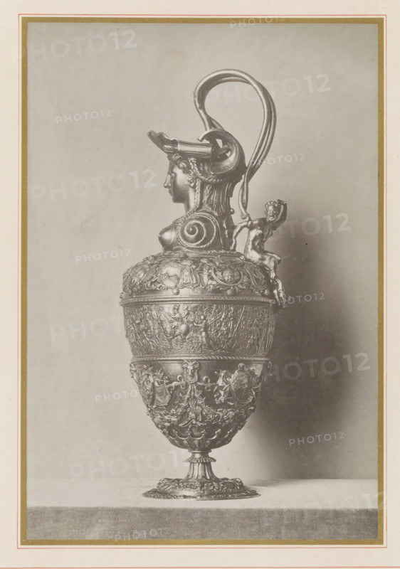 Lamppot of Emperor Charles V, seen from the side, Aiguière de Charles V (profile), Musée National du Louvre Galerie d'Apollon, page, photomechanical print, inscription: 'ateliers de reproductions artistiques', photographer: Léon Vidal, Paris, cliché maker: Léon Vidal, c. 1876 - c. 1883, Léon Vidal, photographer, cliché maker, paper, collotype, phototypie, height 260 mm x width 198 mm, container of precious metal, chasing, embossing (industrial process), A tall ornate metal vessel with a pedestal base sits centered on a flat surface against a plain backdrop, featuring a rounded body densely decorated with relief scenes and decorative motifs, a midsection band with emblematic shields and garlands, and an upper body with a molded lip. The handle rises high above the rim in a looping curve and attaches to a sculpted female head with braided hair and a helmet-like headdress that forms part of the spout area, while a small seated figure clings to the handle near the vessel's shoulder.