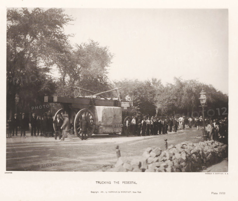 Trucking the pedestal, page, photomechanical print, inscription: 'Copyright 1881, by HARROUN & BIERSTADT, New York', number: 'Plate XVIII', New York, cliché maker: Harroun & Bierstadt (possibly), 1880, photographer, printer (person), North America, paper, collotype, artotype, height 189 mm x width 255 mm, (farm) wagon, freight wagon, cart, obelisk, needle, base (, column, pillar), A large wooden wagon with oversized wooden wheels stands in a tree-lined street, its flatbed carrying a block-shaped stone pedestal and long timber beams with ropes and chains draped over them; a crowd of men in dark suits and hats stands around and alongside the wagon, with some men positioned near the wheels and others in a line behind it, while horses and additional figures are visible further back; tall gas-style street lamps rise near the roadside and a low stone wall runs along the foreground, with leafy trees forming a backdrop and open sky filling the upper part of the image.
