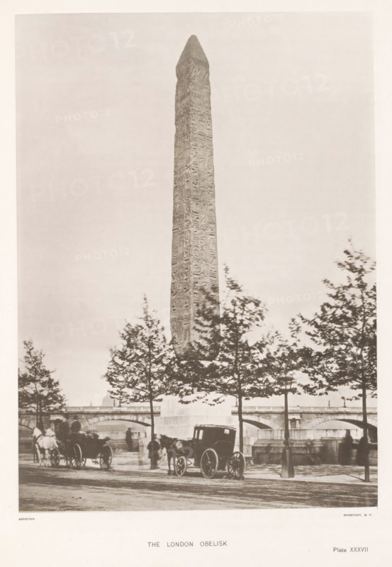 The London obelisk, page, photomechanical print, number: 'Plate XXXVII', cliché maker: Harroun & Bierstadt (possibly), New York (city), c. 1881, photographer, printer (person), North America, paper, collotype, artotype, height 266 mm x width 192 mm, obelisk, needle, London, Cleopatra's needle (London), A tall stone obelisk with carved surface rises vertically from a base and dominates the scene, framed by a row of trees with leafy branches in the foreground; below the obelisk a bridge with arches spans the background and there are horse-drawn carriages and figures on a paved street near the curb, with a silhouette of a cityscape visible behind the bridge.
