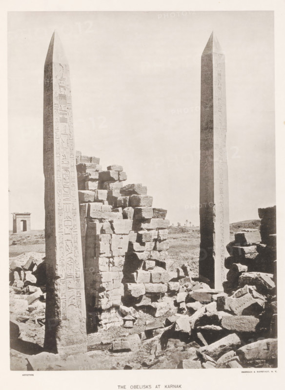 The obelisks at Karnak, page, photomechanical print, number: 'Plate XL', Karnak, cliché maker: Harroun & Bierstadt (possibly), New York (city), c. 1881, photographer, printer (person), North Africa, North America, paper, collotype, artotype, height 274 mm x width 205 mm, ruin of a building, architecture, obelisk, needle, temple, shrine, Egyptian religion, Two tall obelisks rise vertically from a rubble-strewn foreground, each with smooth tapering sides and pointed tops, their surfaces marked with vertical carved designs. Between and around the obelisks are piles of large, rough-hewn stone blocks stacked and scattered, creating irregular shapes and shadows. In the distant background a small rectangular stone structure with an open doorway stands on the plain, and a few thin palm trees are visible near the horizon. The sky above is pale and empty, filling most of the upper part of the image.