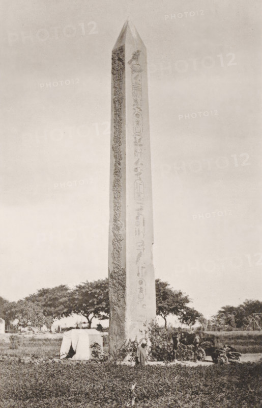 The obelisk at Heliopolis and Pompey's pillar at Alexandria, page, photomechanical print, number: 'Plate XLI', cliché maker: Harroun & Bierstadt (possibly), printer (person): Harroun & Bierstadt, New York (city), c. 1881, Harroun & Bierstadt, photographer, cliché maker, printer (person), North America, paper, collotype, artotype, height 270 mm x width 352 mm, Prints opposite page 124., column, pillar, architecture, obelisk, needle, North Africa, Alexandria, Pompey's Pillar, Serapeum, A tall stone obelisk occupies the right side of the image, showing a pointed top and faint carved markings along its shaft, while the left side shows a grassy field with scattered trees and a group of people and animals near the foreground, all beneath a pale sky.