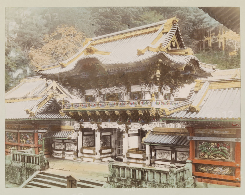 View of the Yomeimon Gate at Nikko, photograph, page, Nikko, Japan, c. 1887 - in or before 1897, photographer, colorer (graphic design artist), Kanto, paper, albumen print, hand coloring, height 186 mm x width 243 mm, Photo for page 63., facade (of house or building), gate, entrance, A large ornate traditional building with layered curved roofs topped with decorative ridge elements and gold-colored accents, intricate carved and painted wooden panels beneath the eaves, and sculpted brackets and ornamental details along the facade; stone steps and a weathered stone balustrade lead up to the entrance area, and painted panels with floral and nature motifs appear on the lower walls; trees and foliage form a background behind the structure.