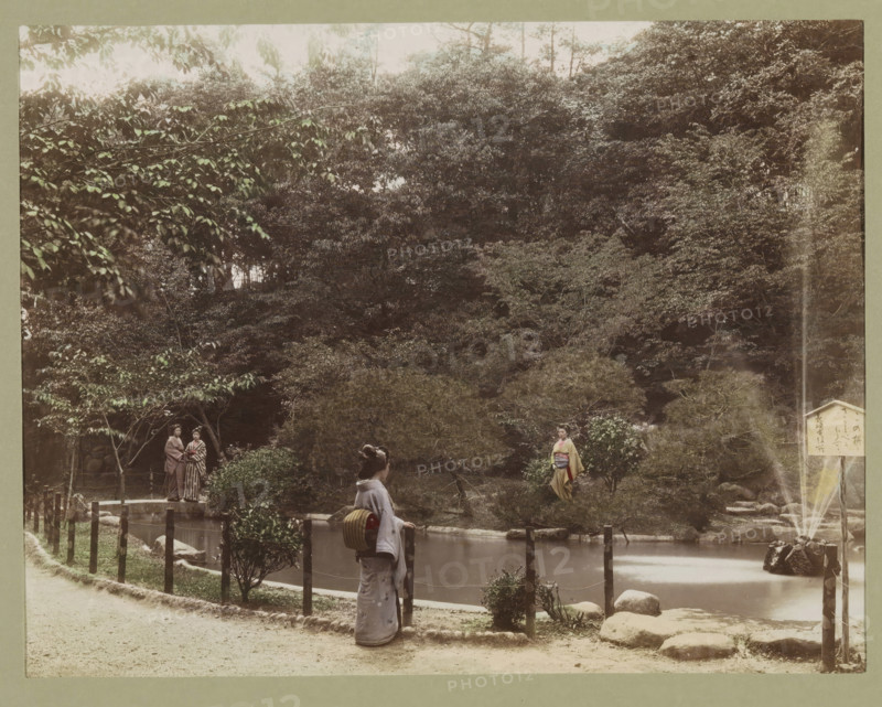 View of a pond in Osuwa Park in Nagasaki, photograph, page, Nagasaki, Japan, c. 1887 - in or before 1897, photographer, colorer (graphic design artist), Kyushu, paper, albumen print, hand coloring, height 188 mm x width 243 mm, Photo for page 135., public gardens, park, pond, pool, A landscaped scene with dense trees and shrubs in the background and a calm pond in the middle foreground, a narrow dirt path running along the front edge, wooden posts and rope forming a low fence beside the pond, a stone fountain sending a vertical stream of water into the pond near a cluster of rocks, a woman in traditional dress standing at the fence facing the water with a bundled item on her back, another woman in traditional dress standing on the far side of the pond among the bushes, and a pair of people standing together near a small bridge partially visible at one side.