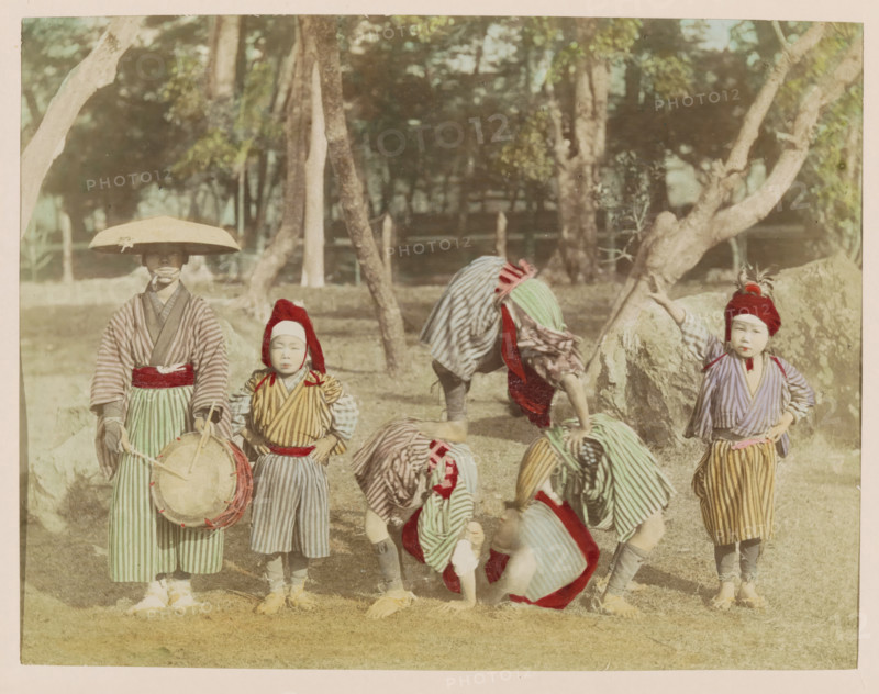 Child acrobats and their attendant in a park, photograph, page, Japan, c. 1887 - in or before 1897, photographer, colorer (graphic design artist), paper, albumen print, hand coloring, height 188 mm x width 243 mm, Photo for page 141., park, acrobat, circus, drum (musical instrument), Several children stand and bend on grass in front of trees and rocks, wearing striped robes and sashes in muted colors, some with red head coverings and one with a wide brim hat, one child holding a round drum with straps, others bent over or crouching with patterned cloths draped over them, and a child standing with a hand on a rock while another child stands with hands at their sides.