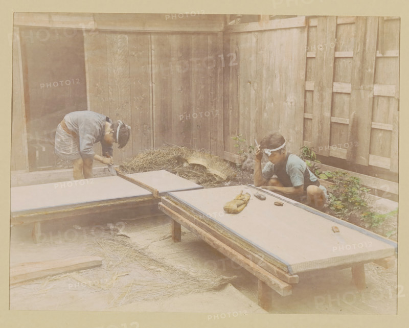 Two mat makers at work, photograph, page, Japan, c. 1887 - in or before 1897, photographer, colorer (graphic design artist), paper, albumen print, hand coloring, height 187 mm x width 243 mm, Photo for page 147., handicrafts, craftsman at work, door furniture, eg boot scraper, brush, doormat, A faded photograph shows two people working in a wooden courtyard with vertical plank walls. One person on the left is bent over a low platform, wearing a light-colored top and short skirt, with a cloth tied around their head. The other person on the right is seated on the ground near a corner of the enclosure, wearing a short-sleeved patterned top and a headband, holding something near their head. Several low wooden tables or boards are arranged across the foreground, one with a small bundle and a few small tools or objects on it. A pile of straw or dried grass rests against the wall between the two people, and some small plants grow near the seated person. The overall scene has muted, sepia-tone...
